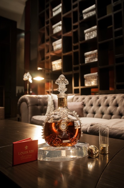 Louis XIII cognac in a flattened round clear-glass decanter with projecting glass fins, embossed medallions and a fleur-de-lis stopper, photographed on a table in a bar.
