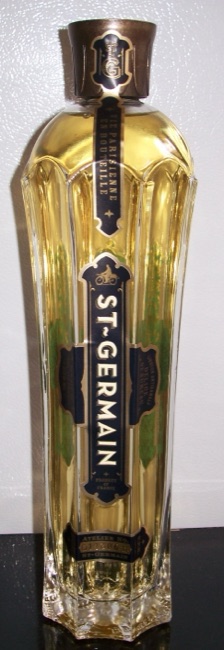 Full tall St-Germain elderflower liqueur bottle with vertical fluted facets, arched shoulders, broad cap and a geometric heavy glass base against a pale wall.