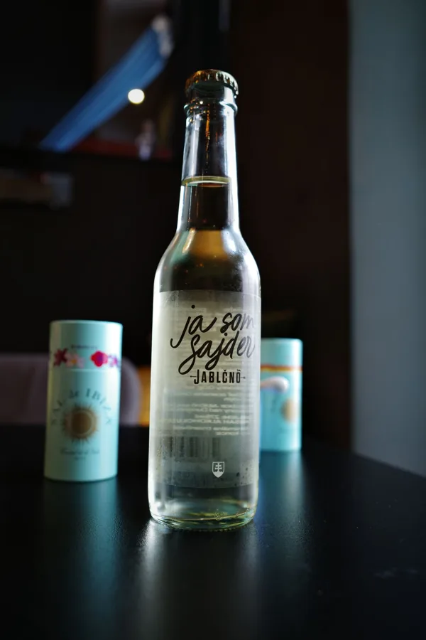 Full clear cider bottle and crown cap on dark table, moody side lighting and shallow background; Ja som Sajder/Jablcno label readable.