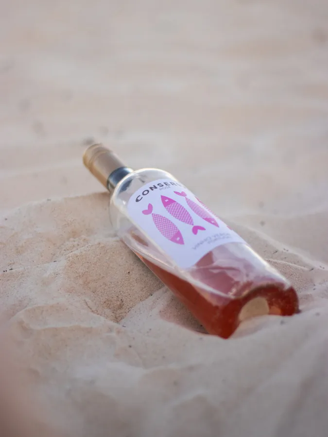 Rose bottle lying diagonally in pale sand; portrait, soft summery palette. Bottle complete but part of lower edge sits in sand. Fish-pattern label and partial Conser... name readable.