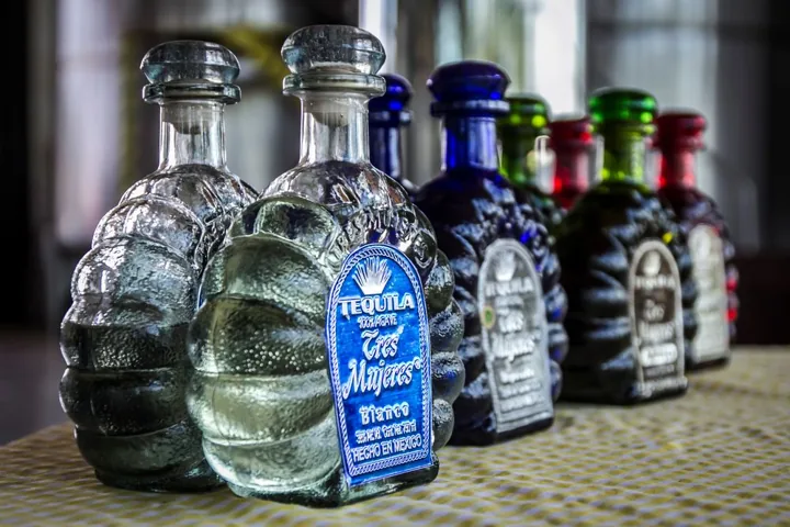 Small row of sculpted, lobed Tres Mujeres tequila bottles with glass stoppers, in clear, cobalt blue, green and red.