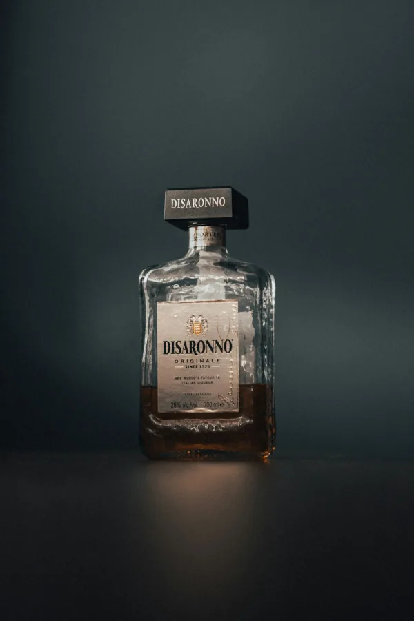 Disaronno liqueur bottle with rectangular body, textured glass and oversized square black cap, muted teal background.